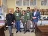 Pięciu pracowników naukowych stoi ramię w ramię w jasnej pracowni uniwersyteckiej, otoczonej próbkami drewna i sprzętem laboratoryjnym. Trzy osoby w centrum mają na sobie zielone bluzy z kapturem z logo „University of Forestry”, natomiast mężczyźni po bokach ubrani są w eleganckie marynarki. Tło wypełniają liczne eksponaty dendrologiczne ułożone na półkach oraz tablice z przekrojami pni drzew zawieszone na ścianie. Cała grupa uśmiecha się w stronę obiektywu.