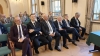 Grupa mężczyzn w garniturach siedzi w pierwszym rzędzie widowni podczas oficjalnego wydarzenia. Większość z nich ma splecione dłonie i patrzy przed siebie z uwagą. W tle, w kolejnych rzędach, widać pozostałych uczestników spotkania, w tym osoby w mundurach leśnika.