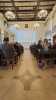 Perspektywa z tyłu sali konferencyjnej, ukazująca rzędy uczestników siedzących na ciemnozielonych krzesłach. Na dużym ekranie z przodu wyświetlane jest zdjęcie prof. Andrzeja Grzywacza z podpisem. Wnętrze charakteryzuje się ozdobnym, kasetonowym sufitem i wiszącymi kryształowymi żyrandolami.