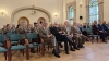 Uczestnicy konferencji siedzą w rzędach, a wielu z nich ubranych jest w mundury wyjściowe Lasów Państwowych. W centralnej części kadru siedzi mężczyzna w ciemnej marynarce i okularach, trzymający notatnik. Pomieszczenie jest jasne, z dużymi łukowatymi oknami i dekoracyjną wnęką w ścianie.