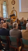 Kobieta w ciemnym stroju przemawia do mikrofonu, stojąc za drewnianą mównicą na tle jasnej ściany. Obok niej stoi mężczyzna w mundurze leśnika, trzymający dokumenty. Na pierwszym planie widoczne są plecy i głowy słuchaczy siedzących w zielonych fotelach, skierowanych w stronę prelegentki.