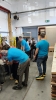 Grupa osób pracuje w przestronnym warsztacie przy montażu drewnianych elementów. Kilku mężczyzn w niebieskich koszulkach nadzoruje pracę młodzieży skupionej wokół stołów roboczych. W pomieszczeniu widoczne są stosy tarcicy, narzędzia oraz konstrukcja drewnianego mostu kratownicowego.