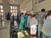 Grupa młodzieży w ochronnych fartuchach stoi przy długim stole laboratoryjnym, obserwując przeprowadzane doświadczenia. Uczniowie są zwróceni tyłem, skupieni na pracy przy stanowiskach z odczynnikami. Nad nimi, na ścianie z białych płytek, wisi fragment tablicy z układem okresowym pierwiastków.