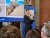 Mężczyzna w garniturze prowadzi wykład, gestykulując ręką i mówiąc do mikrofonu. W tle widoczny jest fragment prezentacji multimedialnej przedstawiającej żółtą ładowarkę pracującą przy hałdzie trocin. Spotkanie ma charakter oficjalnej konferencji lub panelu edukacyjnego.