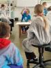 Grupa młodzieży w ochronnych fartuchach stoi tyłem, obserwując pokazy chemiczne w jasnym laboratorium. Uczniowie są skupieni na czynnościach wykonywanych przy stole laboratoryjnym, na którym znajdują się liczne szklane naczynia i odczynniki. W tle widać kafelkowane ściany, tablicę z układem okresowym pierwiastków oraz duże okna wpuszczające naturalne światło do pracowni.