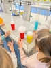 Kobieta w białym fartuchu laboratoryjnym nalewa bezbarwny płyn z kolby do wysokiego cylindra miarowego, w którym znajduje się intensywnie niebieska ciecz. Doświadczenie odbywa się na profesjonalnym stanowisku laboratoryjnym wyposażonym w krany z dostępem do mediów i liczne butelki z odczynnikami w tle. Wokół stołu stoją zaciekawione dzieci, które z bliska obserwują reakcję chemiczną.