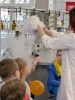 Kobieta w białym fartuchu laboratoryjnym pokazuje dzieciom widowiskowe doświadczenie z użyciem suchego lodu. Z kolb ustawionych na stole wydobywają się kłęby gęstej, białej pary, która opada na blat i przelewa się przez krawędzie naczyń. Uczniowie z bliska obserwują proces, a w tle widoczne są szafki laboratoryjne oraz profesjonalny sprzęt chemiczny.