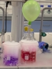Zbliżenie na naczynia laboratoryjne wypełnione kolorowymi płynami, z których wydobywa się gęsta, biała para. Na szczycie jednej ze szklanych butli zamocowany jest jasnozielony, nadmuchany balon. Efektowne doświadczenie wykorzystuje prawdopodobnie suchy lód, tworząc dynamiczną scenę naukową.