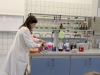 Kobieta w fartuchu laboratoryjnym przygotowuje odczynniki na stanowisku badawczym, pochylając się nad kolorowymi roztworami. Na blacie obok niej stoją zlewki z fioletową i czerwoną cieczą oraz naczynia z parującym białym dymem. W tle, na regałach, poukładane są liczne butelki z chemikaliami opatrzone etykietami.