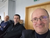 Trzej mężczyźni siedzą rzędem podczas spotkania w jasnym pomieszczeniu. Na pierwszym planie widać uśmiechniętego mężczyznę w okularach i ciemnej marynarce, który wykonuje autoportret. W tle, na dalszym planie, siedzą dwaj pozostali uczestnicy, uważnie patrząc przed siebie.