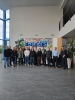 Grupa kilkunastu osób pozuje do wspólnego zdjęcia w wysokim, nowoczesnym lobby z dużą ilością przeszkleń. Nad nimi wisi kolorowy, ręcznie malowany transparent z logo firmy Remmers. Uczestnicy stoją swobodnie na szarej, błyszczącej posadzce, a obok nich widoczne są duże rośliny doniczkowe.