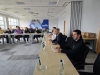 Widok z boku na uczestników siedzących przy długim stole podczas spotkania biznesowego. Na blacie stoją szklanki oraz butelki z wodą mineralną. W tle widać dużą fototapetę z logo firmy oraz postacie siedzące po drugiej stronie sali.