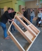 Grupa mężczyzn w ciemnych bluzach i jeansach wspólnie podnosi oraz obraca dużą, przestronną konstrukcję z drewnianych belek i płyt OSB. Pracy z boku przyglądają się dwie studentki stojące w pobliżu otwartych drzwi prowadzących do kolejnego pomieszczenia. Zajęcia praktyczne odbywają się na betonowej posadzce wewnątrz przestronnego warsztatu stolarskiego lub budowlanego.