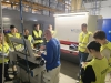 Instruktor w niebieskiej bluzie z logo Sherwin-Williams demonstruje obsługę specjalistycznej maszyny przemysłowej, regulując jej elementy na oczach zebranej młodzieży. Wokół niego stoi grupa studentów w żółtych kamizelkach odblaskowych, którzy z bliska obserwują cały proces technologiczny. W tle wyróżnia się duże, jasne urządzenie marki Weber z podświetleniem oraz wysokie regały magazynowe wypełnione towarami. Cała prezentacja odbywa się w dobrze oświetlonym, nowoczesnym otoczeniu hali produkcyjnej.