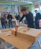Młody mężczyzna w czarnej bluzce z długim rękawem precyzyjnie przycina piłą ręczną gruby blok jasnobrązowej izolacji termicznej. Materiał spoczywa na drewnianej płycie opartej na kobyłkach warsztatowych, a obok leży małe, czerwono-białe pudełko. Pracy z uwagą przygląda się grupa studentów oraz instruktor, stojący w tle jasnej sali szkoleniowej. W otoczeniu warsztatu widoczne są makiety konstrukcji dachowych oraz tablice poglądowe z rysunkami technicznymi.