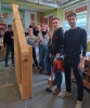 Grupa uśmiechniętych studentów pozuje do wspólnego zdjęcia obok gotowej, wysokie konstrukcji drewnianej ściany szkieletowej. Na pierwszym planie dwaj młodzi mężczyźni trzymają narzędzia budowlane: duży młot kowalski na długim trzonku oraz pomarańczową gwoździarkę gazową. Pozostali uczestnicy zajęć stoją w rzędzie za nimi, trzymając w dłoniach miarę zwijaną oraz kątownik stolarski.