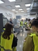 Grupa studentów w żółtych kamizelkach odblaskowych ogląda próbki drewnianych paneli w jasnym laboratorium przemysłowym. Młodzi ludzie trzymają w dłoniach kwadratowe fragmenty materiałów o różnej kolorystyce i strukturze, porównując ich wykończenie. Po lewej stronie brodaty instruktor w okularach i ciemnej kamizelce rozmawia z uczestnikami, gestykulując dłońmi. W tle widoczne jest specjalistyczne wyposażenie techniczne, w tym laboratoryjne stanowiska robocze ze zlewem, rury wentylacyjne pod sufitem oraz plan