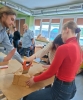Studentka w jaskrawoczerwonym golfie i jeansach stabilnie przytrzymuje na stoliku warsztatowym duży blok jasnobrązowej izolacji termicznej. Tuż obok niej druga osoba precyzyjnie przecina materiał za pomocą piły ręcznej z pomarańczową rękojeścią, a stojący po lewej stronie chłopak asekuruje cały proces dłonią. W tle jasnej sali lekcyjnej, wyposażonej w duże okna i system wentylacji podsufitowej, zajęciom z uwagą przyglądają się pozostali studenci oraz brodaty instruktor.