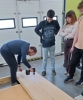 Mężczyzna w niebieskim swetrze, mocno pochylony nad leżącą na posadzce płytą pilśniową marki Steico, wkręca w nią długi wkręt za pomocą akumulatorowej wkrętarki. Jego pracy z uwagą przygląda się stojąca obok trójka studentów, z których dziewczyna ma splecione na piersiach ramiona. Zajęcia odbywają się na gładkiej podłodze warsztatowej na tle zamkniętej, segmentowej bramy garażowej z niewielkimi oknami.