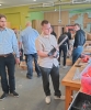 Młody mężczyzna w okularach i jasnej koszuli stoi w centrum warsztatu, uważnie oglądając małą, akumulatorową pilarkę łańcuchową trzymaną w dłoniach. Obok niego po lewej stronie stoi kolega w błękitnej koszuli, podczas gdy w tle pozostali uczestnicy zajęć pracują przy drewnianych konstrukcjach szkieletowych. Po prawej stronie widoczny jest masywny stół stolarski z rozłożonymi narzędziami, takimi jak piła ręczna, młotek oraz pomarańczowe elektronarzędzia.
