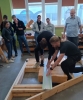 Dwaj mężczyźni na pierwszym planie precyzyjnie przycinają wełnę mineralną przy użyciu metalowej prowadnicy i narzędzia do cięcia. Ich pracy, skupionej na konstrukcji drewnianego szkieletu, przygląda się grupa studentów i instruktor w jasnej sali warsztatowej. Obserwatorzy stoją w tle, zwróceni w stronę demonstrowanej techniki izolacji. Pomieszczenie jest doświetlone naturalnym światłem z dużych okien, a na półkach widać inne projekty i narzędzia.