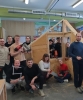 Grupa uśmiechniętych młodych ludzi pozuje do wspólnego zdjęcia w warsztacie wokół drewnianej makiety małego domku o konstrukcji szkieletowej. Kilka osób klęczy lub siedzi na podłodze z przodu, a pozostali stoją wokół konstrukcji, niektórzy zaglądając przez otwory okienne. Wiele osób trzyma w rękach narzędzia budowlane i stolarskie, takie jak gwoździarka, piła ręczna, wkrętarka czy rozkładana miara.
