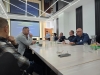 Grupa mężczyzn uczestniczy w spotkaniu biznesowym lub naukowym, siedząc wokół długiego, jasnego stołu konferencyjnego. Jeden z uczestników po prawej stronie wnikliwie analizuje trzymany w dłoni dokument, podczas gdy inni rozmawiają lub słuchają w skupieniu. Na końcu sali widoczny jest duży ekran z prezentacją, a w głębi stoi mężczyzna z kartkami papieru.