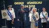 Mężczyzna w garniturze i starsza kobieta w jasnym kostiumie stoją na scenie, trzymając się za uniesione dłonie w geście triumfu. Kobieta ma na ramieniu błękitną szarfę i złoty łańcuch z medalem, a w ręku trzyma mikrofon. Towarzyszą im dwaj chłopcy w białych koszulkach z logiem słońca, z których jeden trzyma proporzec, a drugi czerwoną różę. W tle widnieje duży napis „ORDER UŚMIECHU” oraz data „2.04.2026”.