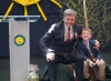 Uśmiechnięty mężczyzna w eleganckim garniturze i chłopiec w podobnym stroju siedzą na pieńkach na scenie. Mężczyzna obejmuje chłopca ramieniem, a w tle widać ozdobę w kształcie słońca oraz brzozowe gałązki. Obok stoi przezroczysta mównica z logiem żółtego słońca. Na podłodze przed nimi leży zafoliowany prezent, a na twarzach obu widać radość.