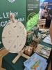 Drewniane koło fortuny z wypalonymi symbolami prezentów i skrótem UPP stanowi główną atrakcję stoiska Wydziału Leśnego i Technologii Drewna. Obok urządzenia, na zielonym obrusie, estetycznie ułożono liczne gadżety promocyjne: ołówki w drewnianych kubkach, breloki, zakładki do książek oraz stosy informacyjnych ulotek. W tle widoczny jest duży, zielony baner z logotypem wydziału oraz fragmenty sąsiednich wystaw targowych.