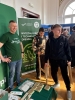 Uśmiechnięty przedstawiciel Wydziału Leśnego i Technologii Drewna UPP, ubrany w zieloną koszulkę promocyjną, stoi obok roll-upu uczelni. Przed nim dwaj młodzi mężczyźni testują stabilność i konstrukcję ekologicznego, drewnianego krzesła, które można samodzielnie złożyć. Na stoliku obok wyeksponowano rzędy notesów z hasłem „Mam UPP w pamięci” oraz inne materiały promocyjne.