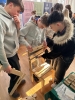 Trzej młodzi mężczyźni wspólnie składają drewniane krzesło wykonane z jasnej sklejki, precyzyjnie dopasowując poszczególne elementy konstrukcyjne. Zadanie wymaga skupienia i współpracy, co przyciąga uwagę przechodniów w gwarnej sali targowej. W tle widoczne są kolorowe banery informujące o kierunkach studiów takich jak kosmetologia, dietetyka oraz stoisko Uniwersytetu Pomorskiego w Słupsku. Całość prezentuje praktyczny aspekt technologii drewna i angażujący charakter warsztatów promocyjnych.