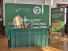 Stoisko promocyjne Wydziału Leśnego i Technologii Drewna Uniwersytetu Przyrodniczego w Poznaniu przygotowane na targi edukacyjne. Na stole przykrytym zielonym obrusem wyeksponowano drewniane koło fortuny, materiały informacyjne oraz gadżety w ekologicznym stylu, takie jak papierowe torby i drewniane kubki z przyborami do pisania. Tło stanowi duża zielona ścianka oraz roll-up z logo uczelni, a obok stoi nowoczesne, jasne krzesło wykonane z drewna.