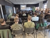 Grupa osób siedzi w rzędach na czarnych krzesłach, przodem do ekranu projekcyjnego wyświetlającego prezentację o przyrodzie Etiopii. Prelegentka prowadzi wykład w nowoczesnej sali, której ściany zdobi leśna fototapeta oraz zielony baner Wydziału Leśnego i Technologii Drewna. Wnętrze jest jasne, z widocznymi drewnianymi regałami pod oknami po prawej stronie. Uczestnicy spotkania są ubrani w codzienne, swobodne stroje i z uwagą śledzą wystąpienie.