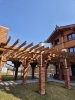 Solidna, drewniana pergola wsparta na ceglanych filarach tworzy geometryczny wzór cieni na trawniku i chodniku. Każdy filar wieńczy ozdobny, bogato rzeźbiony wspornik, który podtrzymuje ciężkie belki konstrukcyjne dachu. W tle widać fragment budynku o spójnej stylistyce, z elewacją wykonaną z drewna układanego w jodełkę oraz dekoracyjnym szczytem dachu. Całość założenia architektonicznego prezentuje się niezwykle rzemieślniczo i estetycznie w pełnym słońcu.