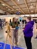 Przedstawicielka uczelni w zielonej koszulce rozmawia z kobietą w fioletowej bluzie na terenie hali targowej. Obok nich stoi drewniane koło fortuny zamocowane na sztaludze, wykorzystywane do konkursów i animacji dla odwiedzających. W tle rozciąga się przestrzeń targowa z widocznymi stoiskami innych wystawców.