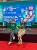 Dwie młode kobiety w zielonych koszulkach siedzą na brzegu sceny, szeroko rozkładając ręce w powitalnym geście. Za nimi znajduje się duży, niebieski ekran multimedialny z napisem „Targi Edukacyjne Poznań 13-15 marca 2026” oraz hasłem „Edukacja w podróży”. Scena jest wyposażona w ciemnozielone fotele i niskie stoliki kawowe, a podest graniczy z czerwonym dywanem na podłodze. Cała kompozycja podkreśla nowoczesny i dynamiczny charakter wydarzenia.