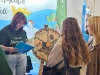Pracowniczka uczelni w zielonej koszulce przegląda niebieski folder informacyjny, stojąc obok dwóch kobiet zainteresowanych ofertą edukacyjną. Pomiędzy nimi znajduje się koło fortuny z widocznymi kategoriami tematycznymi, takimi jak „Alternatywne źródła energii”, „Drzewa i krzewy”, „Gospodarka odpadami” oraz „Grzyby”. W tle widać fragment kolorowej ścianki oraz zarys postaci odwiedzających targi.