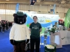 Mascotka uczelniana w stroju bobra z czapką z napisem „UPP” stoi obok młodego mężczyzny w zielonej koszulce na tle stoiska Uniwersytetu Przyrodniczego w Poznaniu. Centralnym punktem tła jest jasna ścianka z hasłem „Przyrodnicze studia” oraz grafikami promującymi nowoczesne rolnictwo i technologię. Na pierwszym planie widać materiały promocyjne rozłożone na białych ladach oraz stojak z ulotkami na tle niebieskiej wykładziny.