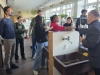 Grupa studentów i wykładowców gromadzi się wokół stanowiska z ciemnymi zlewami w laboratorium, przyglądając się prezentowanym produktom. Mężczyzna w marynarce podaje białe opakowanie kobiecie w różowej bluzie, podczas gdy pozostali uczestnicy z uwagą śledzą proces lub analizują etykiety innych preparatów. Pomieszczenie jest jasne, z dużymi oknami i roślinami doniczkowymi na regałach w tle.