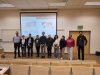 Grupa osób stoi w rzędzie na tle ekranu projekcyjnego w auli wykładowej, prezentującego wykresy sprzedaży i mapę świata firmy Jowat. Uczestnicy znajdują się przed pierwszymi rzędami drewnianych ław studenckich w nowoczesnej, jasnej sali. Po prawej stronie widoczne są drzwi z zielonym znakiem wyjścia ewakuacyjnego.