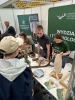 Odwiedzający w beżowej czapce z daszkiem stoi przy stoisku promocyjnym, gdzie dwoje studentów prezentuje materiały edukacyjne związane z naturą. Na stole rozłożone są m.in. poroże jelenia, czaszka dzikiego zwierzęcia, modele maszyn leśnych oraz drewniane koło fortuny. Tło stanowią zielone ścianki reklamowe Uniwersytetu Przyrodniczego w Poznaniu oraz Regionalnej Dyrekcji Lasów Państwowych w Zielonej Górze, z widocznymi trybunami w oddali.