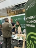 Starszy mężczyzna w oliwkowej kamizelce rozmawia z dwojgiem studentów reprezentujących Wydział Leśny i Technologii Drewna. Na pierwszym planie po prawej stronie widoczny jest fragment zielonego baneru z nazwą wydziału, a na stole leżą ulotki i drewniane koło fortuny. Studenci z uśmiechem udzielają informacji, stojąc na tle zielonej ścianki z logo Uniwersytetu Przyrodniczego w Poznaniu.