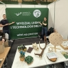 Dwoje studentów pozuje z uśmiechem przy zielonej ściance promocyjnej Wydziału Leśnego i Technologii Drewna Uniwersytetu Przyrodniczego w Poznaniu. Na pierwszym planie, na białym stole, wyeksponowano różnorodne rekwizyty: poroże jelenia, czaszkę zwierzęcia, drewniane koło fortuny oraz mały model maszyny leśnej. Obok nich znajdują się materiały informacyjne, zielone cukierki w drewnianej miseczce oraz dwa miniaturowe krzesła wykonane z drewna.