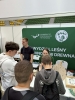 Trójka młodych chłopców stoi plecami do obiektywu, przyglądając się z zainteresowaniem ekspozycji na stoisku Wydziału Leśnego i Technologii Drewna. Za stołem dwoje studentów w skupieniu udziela informacji, prezentując pomoce edukacyjne, w tym drewniane koło fortuny oraz gablotę z owadami. W tle dominuje zielona ścianka z logotypem Uniwersytetu Przyrodniczego w Poznaniu, ustawiona wewnątrz dużej hali z widocznymi rzędami trybun.