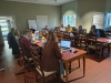 Grupa osób siedzi przy długich, drewnianych stołach ustawionych w kształt litery U w jasnej sali konferencyjnej. Uczestnicy spotkania korzystają z laptopów, robią notatki i prowadzą dyskusję w skupieniu. Na stołach widoczne są kubki z napojami, kolorowe karteczki samoprzylepne oraz materiały szkoleniowe. W tle znajdują się białe tablice typu flipchart oraz klasyczne drzwi i okna, przez które wpada naturalne światło.