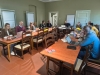 Uczestnicy warsztatów siedzą przy dwóch rzędach drewnianych stołów ustawionych w jasnej, klasycznej sali. Większość osób pracuje na laptopach, a na pierwszym planie widoczne są stosy kolorowych karteczek samoprzylepnych i notatnik. W tle znajdują się flipcharty oraz białe drzwi, a cała grupa sprawia wrażenie zaangażowanej we wspólną dyskusję i zadania projektowe.