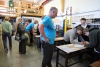 Dwie osoby precyzyjnie przycinają wąską drewnianą listewkę za pomocą małej piłki ręcznej. Praca odbywa się na blacie roboczym, na którym leżą dokumenty techniczne i inne elementy konstrukcyjne. To zbliżenie podkreśla manualny charakter warsztatów i dbałość o detale wykonywanych modeli.