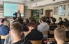 Wykładowca omawia historię szkoły lutniczej w Cremonie, trzymając w rękach korpus skrzypiec. Na ekranie w tle widoczne są nazwiska słynnych mistrzów, takich jak Stradivari, oraz zdjęcie włoskiej kamienicy. Uczniowie skupieni w ławkach przyglądają się prezentacji, a w dolnym rogu kadru widać czerwoną gitarę trzymaną przez jednego z uczestników.
