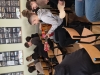Grupa młodzieży w ławkach ogląda z bliska rekwizyty przyniesione na zajęcia – chłopak w szarej bluzie trzyma gitarę akustyczną, a jego kolega obok przygląda się surowej płycie wierzchniej instrumentu. W tle inni uczniowie rozmawiają i oglądają tablice ze zdjęciami zawieszone na ścianie. Atmosfera spotkania sprzyja bezpośredniemu kontaktowi z omawianymi przedmiotami.